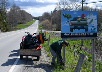 Photo of a man picking up litter along the side of a road with an anti littering sign behind him.