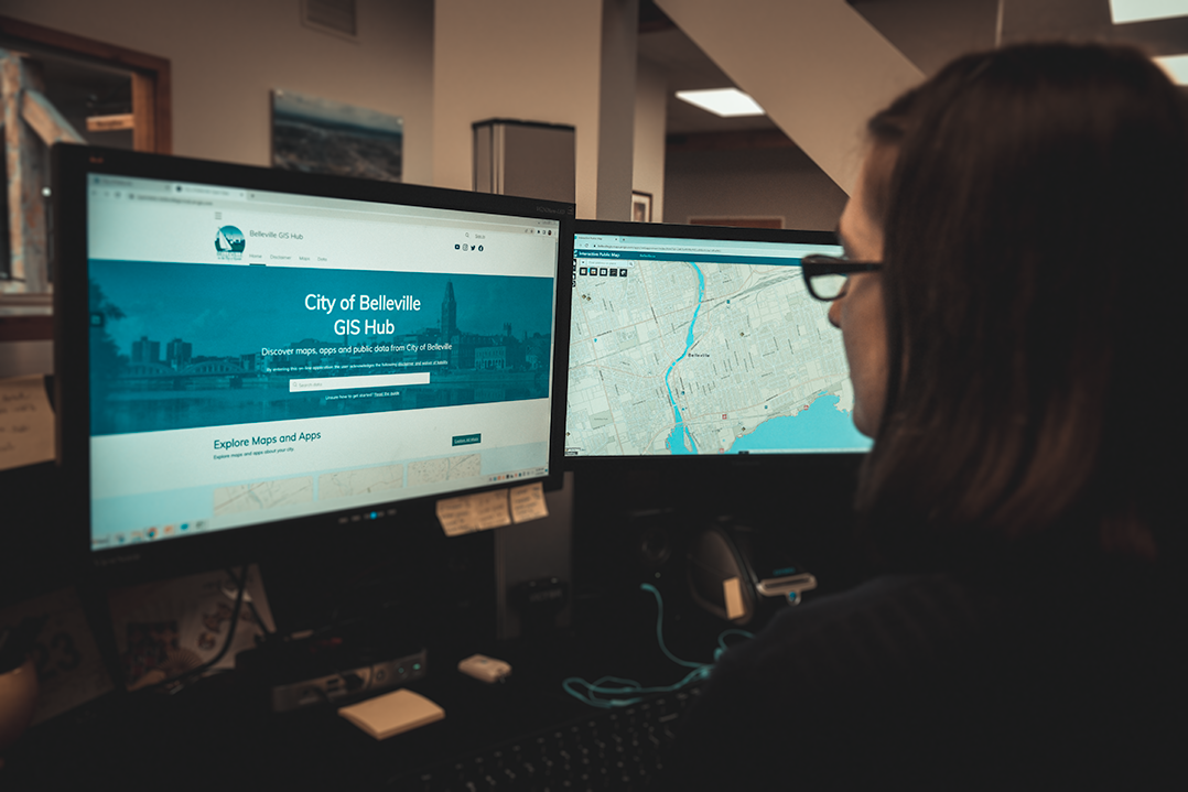 Woman looks at a computer monitor showing the GIS Hub with a second monitor showing a map in the background