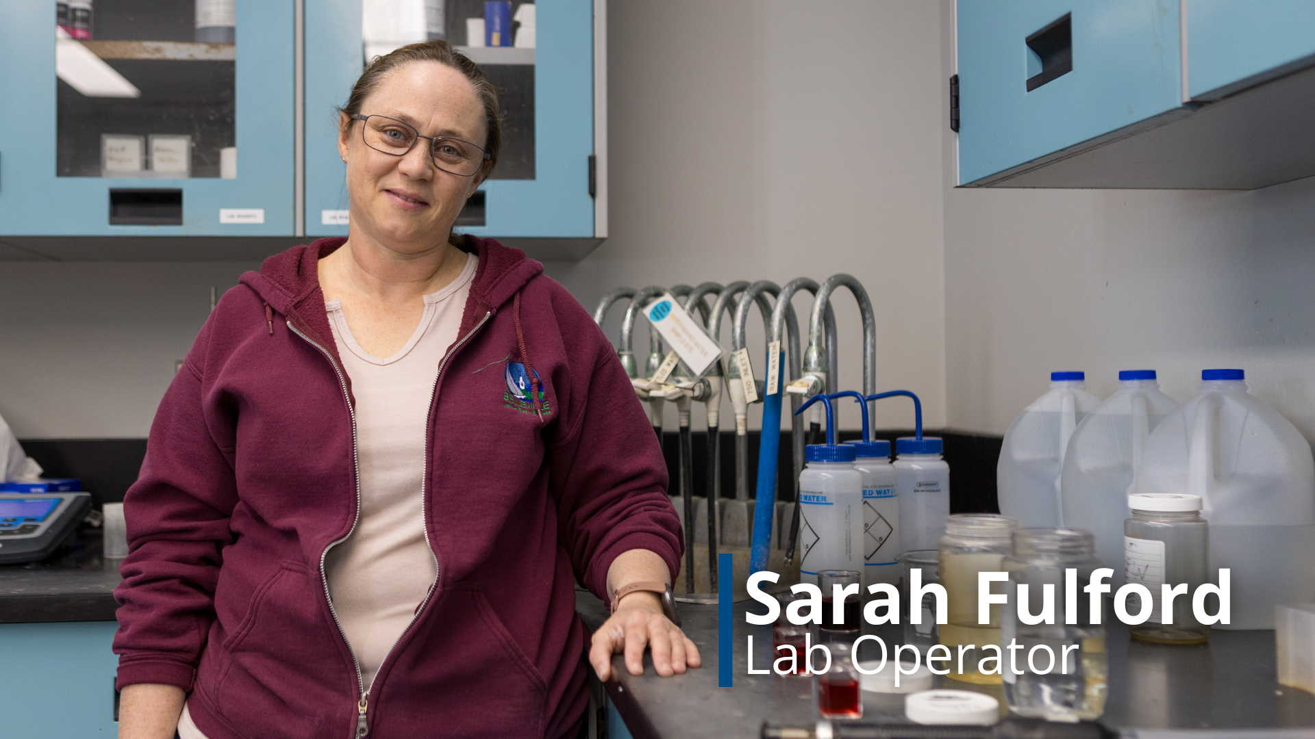 A photo of Sarah Fulford, a Lab Operator, leaning again a counter with beaker and equipment used to test water samples.
