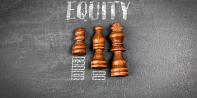Wooden chess pieces of different heights aligned on a chalkboard, with the word “EQUITY” written above them, symbolizing fairness and equal opportunity.