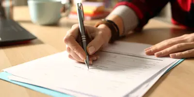 A person filling out and signing a paper form at a desk, with a laptop and coffee cup nearby.