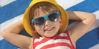 Child laying with arms folded behind head on a beach towel with sunglasses and beach hat